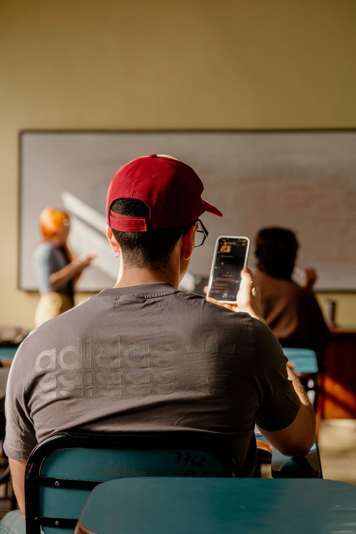 Student sitting in a classroom using a smartphone while the teacher conducts the lecture.